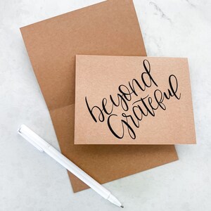 May include: A brown card with the text "beyond grateful" written in black calligraphy. The card is sitting on a brown envelope with a white pen next to it.