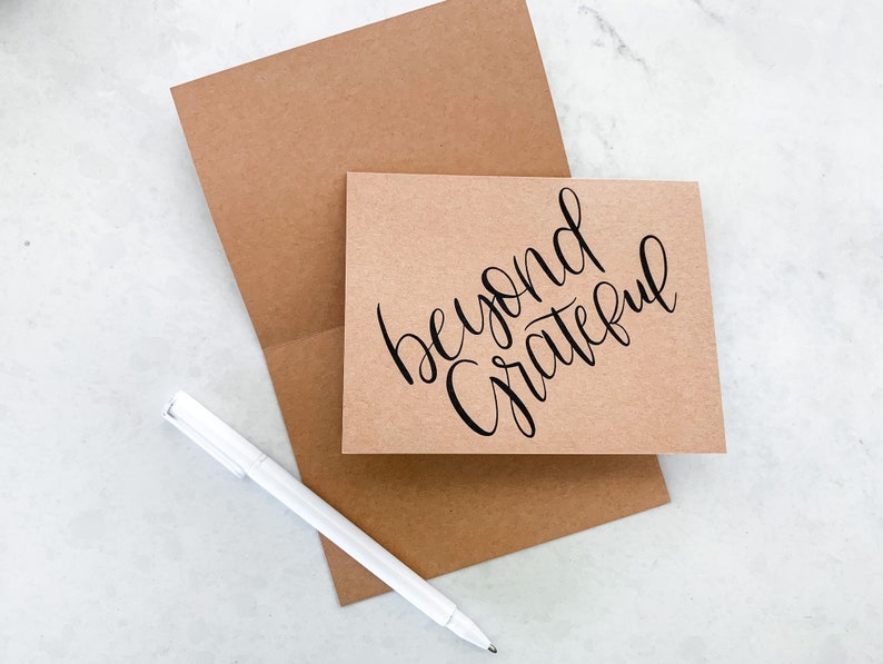 May include: A brown card with the text "beyond grateful" written in black calligraphy. The card is sitting on a brown envelope with a white pen next to it.