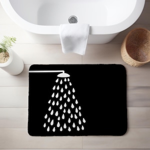 May include: Black rectangular bath mat with a white graphic of a shower head and water droplets. The mat is placed in front of a white bathtub. A white towel hangs on the tub, and a woven basket is nearby.