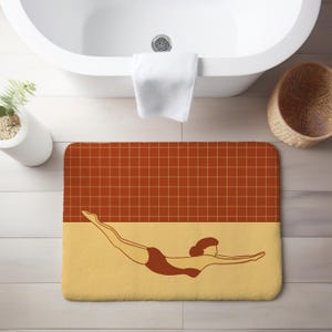 Può includere: Un tappetino da bagno rettangolare con un design retrò. La metà superiore presenta un motivo a griglia di colore marrone rossiccio, mentre la metà inferiore è giallo chiaro. Un nuotatore in costume da bagno marrone scuro è raffigurato nella sezione gialla.