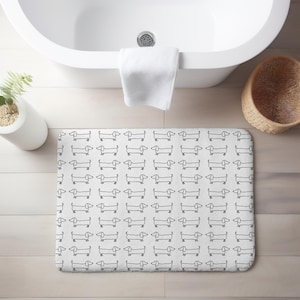 May include: White rectangular bath mat with a repeating pattern of black dachshund dog illustrations. The mat is placed on a light wood floor in front of a white bathtub. A white towel hangs on the tub.
