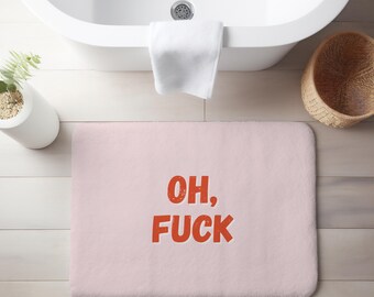 Puede incluir: Una alfombra de baño rectangular de color rosa claro con las palabras "OH, FUCK" en letras naranja desgastadas. La alfombra está colocada frente a una bañera blanca, con una toalla blanca colgada.