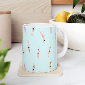 Pode incluir: Caneca de cerâmica branca com fundo azul claro com um padrão de pessoas mergulhando na água. Os mergulhadores são retratados em vários trajes de banho com detalhes coloridos. A caneca está sobre um porta-copos bege.