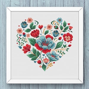 Puede incluir: Un diseño de corazón floral bordado a punto de cruz enmarcado. El corazón está lleno de flores rojas, azules y rosas, hojas verdes y pequeños corazones azules. La obra de arte está en un marco blanco, sobre un fondo de vetas de madera azul claro.