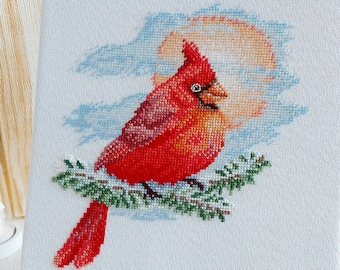 May include: A red cardinal cross-stitch on a white canvas, perched on a branch with green leaves. The cardinal is facing right and has a yellow beak. The background is a blue sky with white clouds.