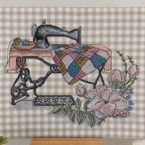 以下が含まれることがあります： ピンクのテーブルとパッチワークのキルトが付いたヴィンテージスタイルのミシン。ミシンは、ベージュのギンガムチェックの背景にピンクと白の花で囲まれています。