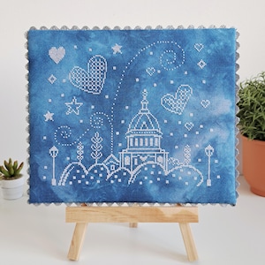 May include: A cross-stitch piece featuring a blue background with white embroidered elements. The design includes a building, hearts, stars, and a scalloped border. The artwork is displayed on a small wooden easel.