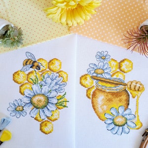 May include: Cross stitch pattern featuring two designs. The first design shows a bee on a honeycomb with white daisies. The second design shows a honey pot with a dipper and white daisies.