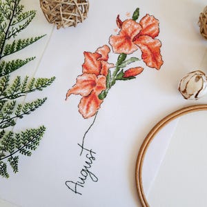 May include: Cross-stitch embroidery of orange flowers with green stems and leaves. The word "August" is written in cursive below the flowers.
