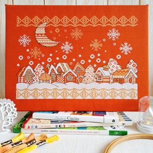 May include: A cross-stitch pattern on orange fabric depicting a winter scene with a crescent moon, snowflakes, and a row of houses. The pattern is framed and displayed on a shelf with books and embroidery floss.
