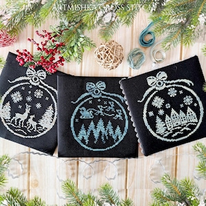 Könnte beinhalten: Drei schwarze Leinwandpaneele mit weißen Kreuzstich-Designs von Weihnachtskugeln. Jede Kugel zeigt eine andere Winterszene: ein Reh, einen Schlitten mit Rentieren und ein Dorf.