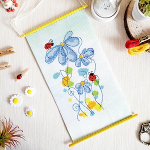 May include: A cross-stitch embroidery of blue flowers with yellow centers and red ladybugs on a white background. The embroidery is framed with a yellow wooden dowel.