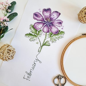 Puede incluir: Patrón de punto de cruz de una flor morada con hojas verdes y la palabra "Febrero" escrita en cursiva debajo.