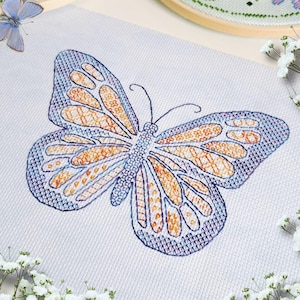 Peut inclure: Broderie au point de croix d'un papillon aux couleurs bleu et orange. Le papillon est au centre, sur un tissu blanc, avec des motifs détaillés dans ses ailes. Des fleurs blanches et un cercle à broder en bois entourent le motif.