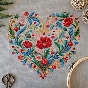 May include: A heart-shaped cross-stitch design featuring red, pink, and blue flowers, green leaves, and gold accents. The floral pattern is set against a gray fabric background. The design includes small red hearts and is surrounded by a wooden embroidery hoop.