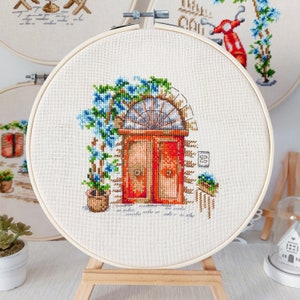 May include: Cross-stitch embroidery hoop with a design of a red door with a blue flower vine and a potted plant. The door has a white frame and a white archway above it. The background is a white canvas.
