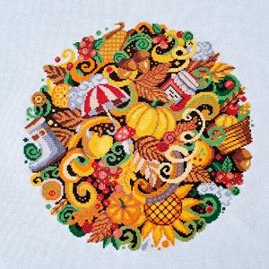 Könnte beinhalten: Ein kreisförmiges Kreuzstichdesign mit Herbstmotiven. Das Design umfasst einen großen gelben Kürbis, einen rot-weißen Regenschirm, eine Sonnenblume und verschiedene Blätter und Beeren in Orange-, Gelb- und Grüntönen. Der Hintergrund ist weiß.
