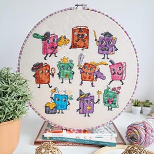 May include: A cross-stitch embroidery hoop with a playful design of cartoon books, each with unique accessories. The books are in various colours, such as pink, orange, green, and purple. The hoop has a striped border and is displayed on a stack of books.