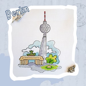 Könnte beinhalten: Ein Kreuzstich-Design mit dem Berliner Fernsehturm, einer Bank und zwei Bäumen. Der Turm ist grau mit einer rot-weißen Antenne. Die Bank ist braun und die Bäume sind grün. Der Hintergrund ist ein hellblauer Aquarell-Wasch.