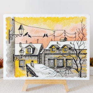 Peut inclure: Une broderie au point de croix représentant un paysage urbain enneigé avec un coucher de soleil jaune et orange. La scène présente plusieurs bâtiments avec des toits et des fenêtres recouverts de neige, un arbre dénudé et des oiseaux perchés sur des lignes électriques.