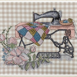 May include: A vintage sewing machine with a pink table and a patchwork quilt on top. The machine is surrounded by a floral arrangement with pink flowers and green leaves.