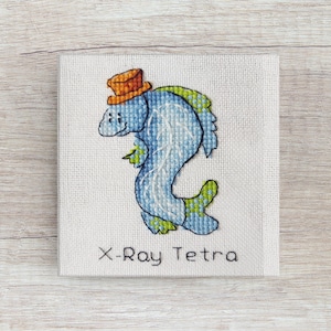Puede incluir: Una bordado de punto de cruz de un pez azul con un sombrero naranja. El pez está etiquetado como "X-Ray Tetra".