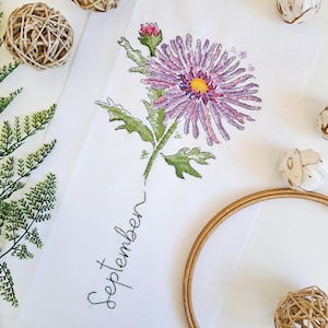 May include: Embroidered design of a purple aster flower with green leaves and stem. The word "September" is embroidered below the flower.