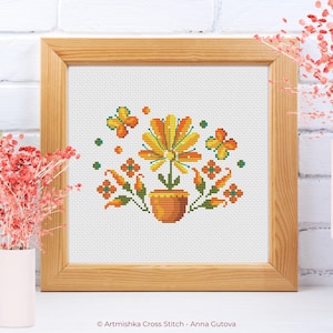 May include: Cross stitch pattern of a potted flower with orange, yellow, and green flowers and leaves. Two butterflies are flying around the flower. The pattern is framed in a wooden frame.