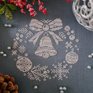 Op de afbeelding: Een kruissteekontwerp met een bel met een strik, omringd door sneeuwvlokken en ornamenten. Het ontwerp is in tinten wit en lichtpaars op een donkergrijze achtergrond. De afbeelding heeft een feestelijk, vakantiethema.