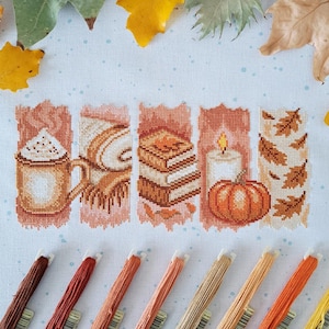 May include: A cross-stitch embroidery pattern on white fabric featuring five vertical panels with autumn motifs: a mug of coffee, a rolled blanket, a stack of books, a lit candle with a pumpkin, and scattered brown leaves, with embroidery floss.