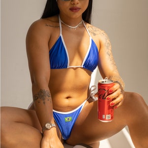 May include: A woman wearing a blue bikini with white trim and a Brazilian flag on the bottom. She is wearing a white headband and sunglasses and holding a red can of soda.