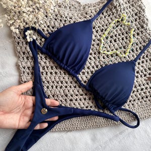 May include: Navy blue bikini set with a triangle top and thong-style bottoms. The bikini is displayed on a beige crocheted bag, with a delicate yellow and white beaded necklace. A small gold-colored button is on the bottom.