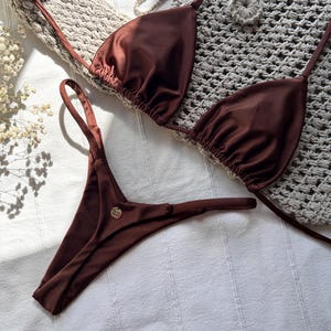 Może przedstawiać: Brązowy zestaw bikini z trójkątnym topem i stringami. Bikini wykonane jest z błyszczącej tkaniny i ma cienkie ramiączka. Mały, złoty emblemat znajduje się na dole. Bikini jest prezentowane na białej powierzchni.