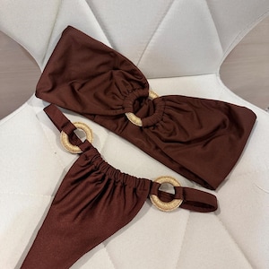 Può includere: Un set di costumi da bagno a due pezzi marrone. La parte superiore è uno stile a fascia con un dettaglio ad anello dorato al centro. La parte inferiore è uno stile perizoma con accenti ad anello dorato. Il set è esposto su una sedia bianca.