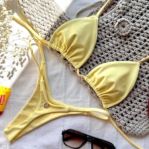 Könnte beinhalten: Ein hellgelbes Bikini-Set mit einem Triangel-Oberteil und String-Hose. Das Bikini liegt auf einer weißen Oberfläche mit einem gehäkelten grauen Cover-up, Sonnenbrille und einer Tube Lippenbalsam.
