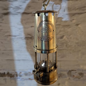 May include: Antique brass miner's safety lamp with a clear glass cylinder and a curved handle. The lamp has a circular emblem with the text "THE PROTECTOR LAMP & LIGHTING CO. 6 M&Q SAFETY LAMPS Approval No. 8/28 MAKERS ECCLES".