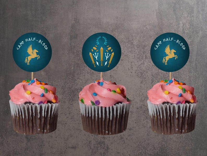 May include: Three cupcakes with pink frosting and colorful sprinkles. Each cupcake has a dark blue topper with the words "CAMP HALF-BLOOD" and a gold illustration of a pegasus or other symbol. The cupcakes are in brown paper liners.