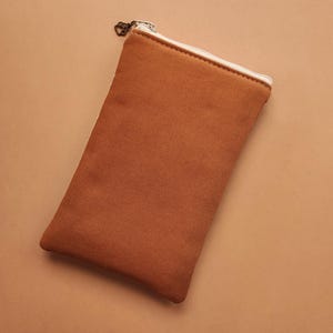 以下が含まれることがあります： 白のジッパーとハート型のジッパープルが付いた、長方形の錆色のキャンバスポーチ。ポーチは閉じられており、お揃いの背景に置かれています。ポーチは、小さなアイテムを保管するために使用される可能性があります。