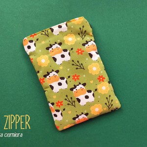 Pu&ograve; includere: Un astuccio rettangolare in tessuto verde con un motivo ripetuto di mucche dei cartoni animati, fiori arancioni e gialli e rametti. Il testo "NO ZIPPER" e "senza cerniera" sono stampati in basso a sinistra.