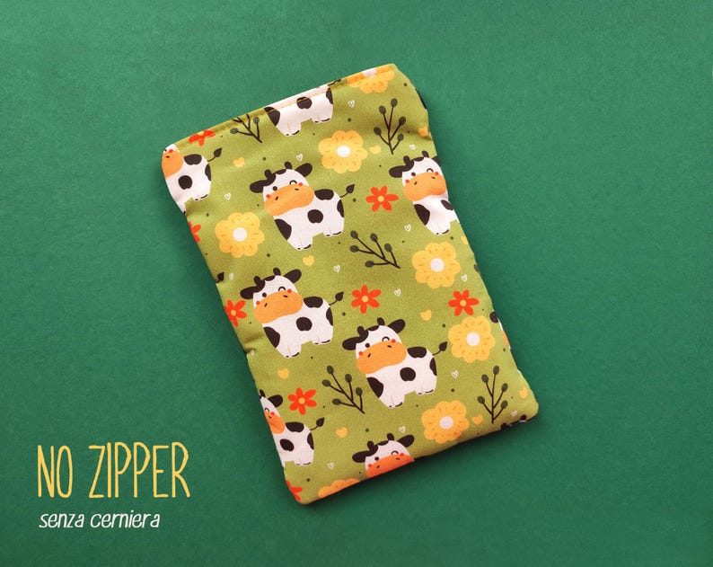 Pu&ograve; includere: Un astuccio rettangolare in tessuto verde con un motivo ripetuto di mucche dei cartoni animati, fiori arancioni e gialli e rametti. Il testo "NO ZIPPER" e "senza cerniera" sono stampati in basso a sinistra.
