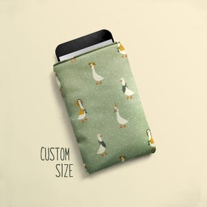 May include: A green phone sleeve with a pattern of geese wearing hats and scarves. The text "CUSTOM SIZE" is printed on the lower left. A black phone is partially visible inside the sleeve.