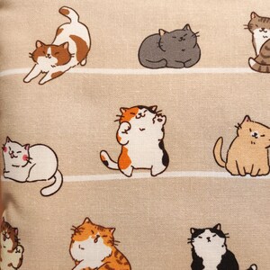 以下が含まれることがあります： さまざまなポーズの漫画の猫の繰り返し模様が特徴のベージュ色の生地。猫は茶色、グレー、オレンジ、黒の色合いです。背景には水平ストライプがあります。