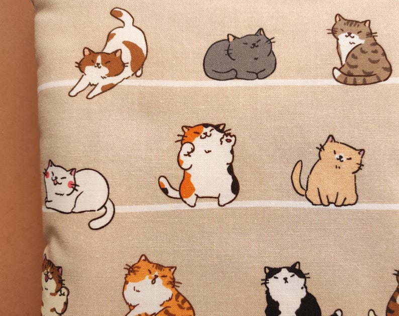 以下が含まれることがあります： さまざまなポーズの漫画の猫の繰り返し模様が特徴のベージュ色の生地。猫は茶色、グレー、オレンジ、黒の色合いです。背景には水平ストライプがあります。