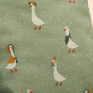 May include: A close-up of a green fabric with a repeating pattern of white geese wearing hats and scarves. The geese have red feet and beaks. The background is a light green with small white dots.