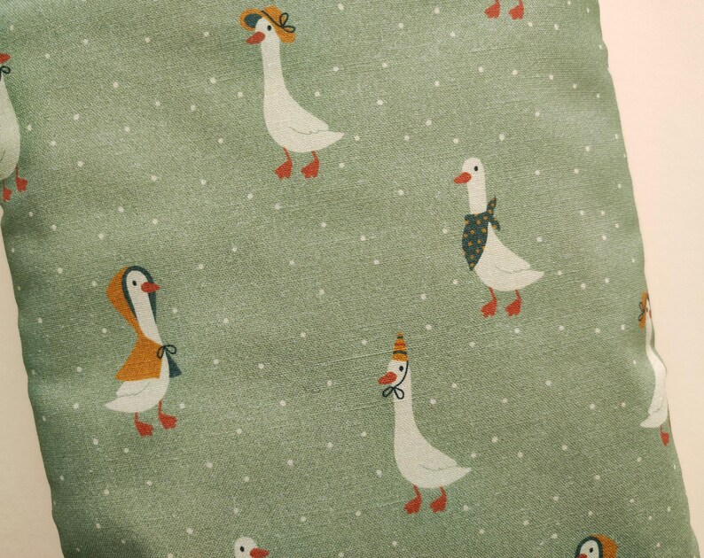 May include: A close-up of a green fabric with a repeating pattern of white geese wearing hats and scarves. The geese have red feet and beaks. The background is a light green with small white dots.