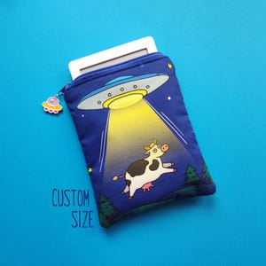 Può includere: Una custodia blu per e-reader con chiusura a cerniera e un design a cartoni animati di un UFO che rapisce una mucca. La custodia ha un piccolo ciondolo sulla cerniera e le parole "CUSTOM SIZE" stampate sul davanti.