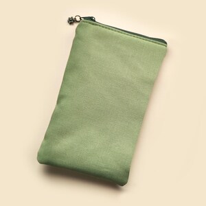 May include: A rectangular, sage green canvas pouch with a dark green zipper and a decorative zipper pull. The pouch is empty and has a simple, minimalist design. The background is a light beige color.