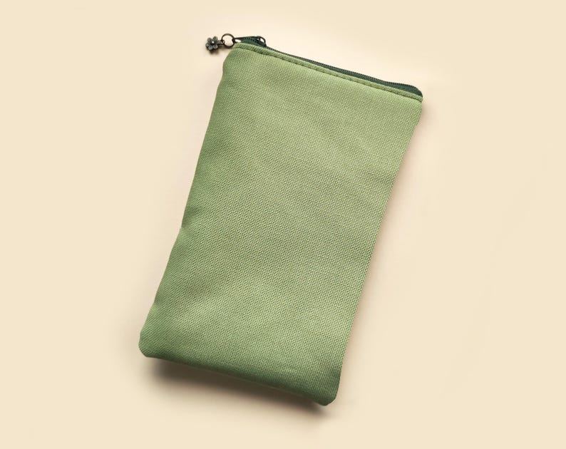 May include: A rectangular, sage green canvas pouch with a dark green zipper and a decorative zipper pull. The pouch is empty and has a simple, minimalist design. The background is a light beige color.