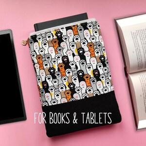 Könnte beinhalten: Eine Reißverschluss-Hülle für Bücher und Tablets mit einem bunten Muster aus Katzenpfoten in verschiedenen Farben. Der untere Teil der Hülle ist schwarz, und der Text "FOR BOOKS & TABLETS" ist auf der Vorderseite aufgedruckt. Ein Tablet und ein offenes Buch sind ebenfalls zu sehen.