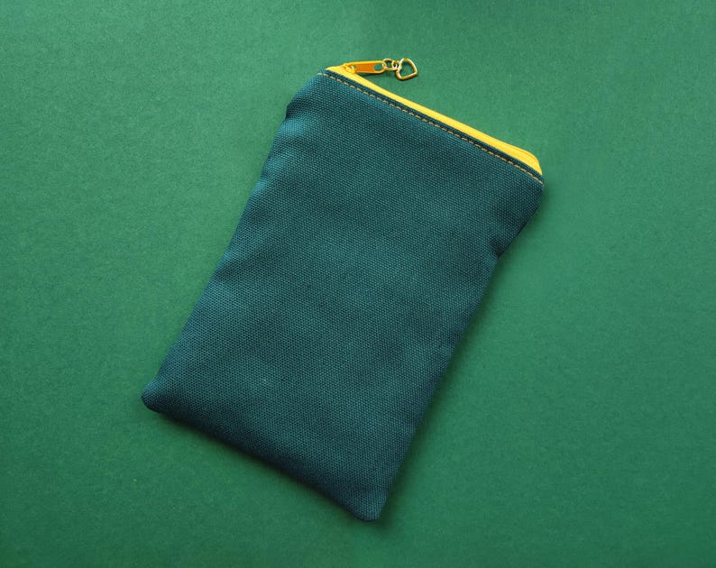 Pu&ograve; includere: Un astuccio in tela verde acqua con cerniera e finiture gialle. L'astuccio rettangolare &egrave; chiuso con una cerniera dorata e poggia su una superficie verde. Design semplice e minimalista.